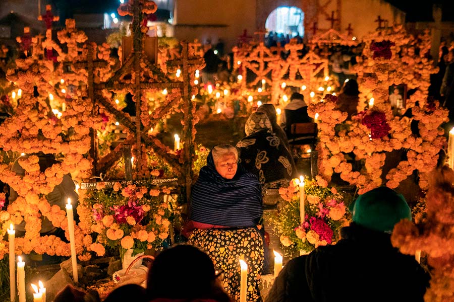 Señora mayor en un cementerio de noche rodeada de velas en celebración por el Día de los Muertos en México.